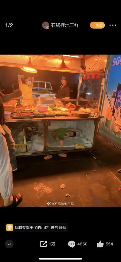 这是一张来自微博的截图，画面呈现了夜晚的街头小吃摊场景：摊位上方悬挂着两盏暖黄色的吊灯，照亮了摆满卤味、小吃的操作台，两位女性摊主在摊位内忙碌，一位抬手整理头发，一位站在一旁。而在小吃摊的金属柜台下方，铺着简易的垫子，一个穿着绿色上衣的小孩正侧躺在上面熟睡，旁边散落着小鞋子和小零食，真实展现了底层劳动者带着孩子辛苦谋生的日常，画面充满了生活的心酸与无奈，引发观者对普通人生活不易的共情。