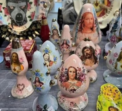 图片展示了一组造型独特的陶瓷工艺品，主要为蛋形或水滴形摆件，表面印有耶稣肖像图案，搭配花卉装饰。这些摆件颜色多样，包括粉色、蓝色等柔和色调，部分作品还带有金属装饰。背景中还可见带有花卉图案和疑似防毒面具图案的盘子等其他陶瓷制品，整体呈现出一种将宗教元素与日常装饰结合的创意风格。