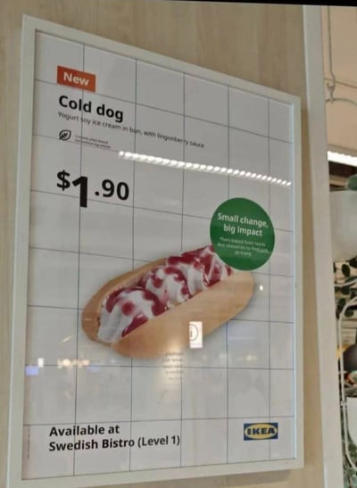 这是一张宜家商场内的新品菜单牌照片，主要宣传名为'Cold dog'的产品。菜单牌背景为白色网格图案，左上角有红色'New'标识，下方是产品名称'Cold dog'及英文描述'Yogurt soy ice cream in bun, with lingonberry sauce'（酸奶大豆冰淇淋放在面包里，配越橘酱）。价格标注为$1.90，中间偏右位置有一张冰淇淋热狗的实物图片，面包中夹着白色冰淇淋和红色酱料。右下角绿色圆形区域写着'Small change, big impact'及'Plant-based food needs less resources to produce, so more'的环保理念文字。底部标注'Available at Swedish Bistro (Level 1)'，右下角有蓝色宜家(IKEA)标志。图片存在双关语：'Cold dog'谐音并对应传统'Hot dog'（热狗），将热食热狗改为冷的冰淇淋版本，形成有趣的产品命名。