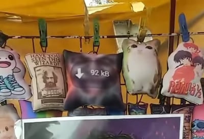 这是一张地摊上卖枕头的照片，展示了多种带有网络和动漫梗的枕头。中间最显眼的是一个写着“↓ 92 kB”的模糊图片梗枕头，模仿低清晰度图片下载感。左侧的英文字母枕头“Have you seen this wizard?”来自哈利波特系列，右侧是猫咪和经典动漫《乱马1/2》的角色图案枕头，还有其他可爱风格的图案。一排枕头通过大夹子夹在钢丝上。整体氛围充满了年轻、搞笑和二次元气息，体现了二创和网络流行文化在现实生活的小商品市场的普及。

特殊注意：“92 kB”枕头是对网络低质量图片的自嘲梗，具有双关和自黑色彩。其设计模仿了图片过度压缩后的模糊感，引发笑点。