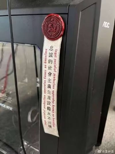 这是一张实拍的黑色电脑主机局部照片，主机为AOC品牌，边框上粘贴了一款趣味装饰贴纸：贴纸顶部是仿红色火漆封印的样式，印有苏式风格的徽章图案，下方连接的仿白色缎带区域竖排印有繁体中文“忠誠的社會主義生產設備永不宕機”，文字两侧还印有对应含义的外文内容，将调侃电脑运行稳定的内容和相关话术结合，制造出诙谐的玩梗效果。