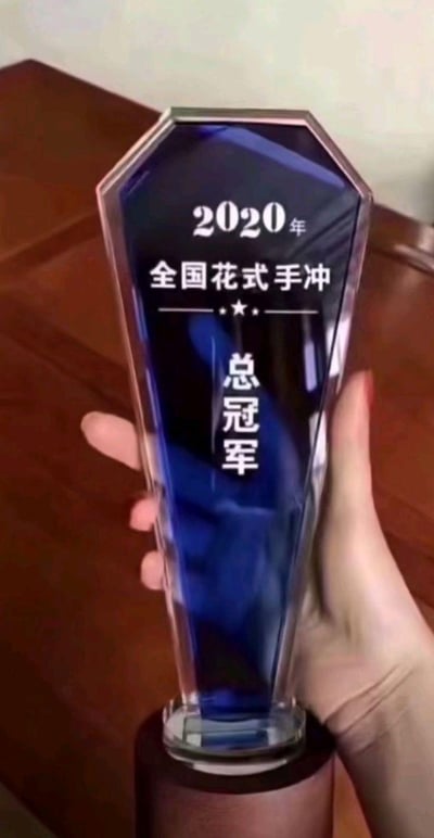 这是一张实拍的荣誉奖杯照片，一只手握着2020年全国花式手冲总冠军奖杯。奖杯主体为蓝透明渐变的水晶材质，顶部带有金属包边，底部是深色木质底座，奖杯表面清晰刻有“2020年”“全国花式手冲”“总冠军”的字样，中间点缀一颗五角星，整体设计精致，彰显了在咖啡手冲赛事中获得的顶级荣誉。