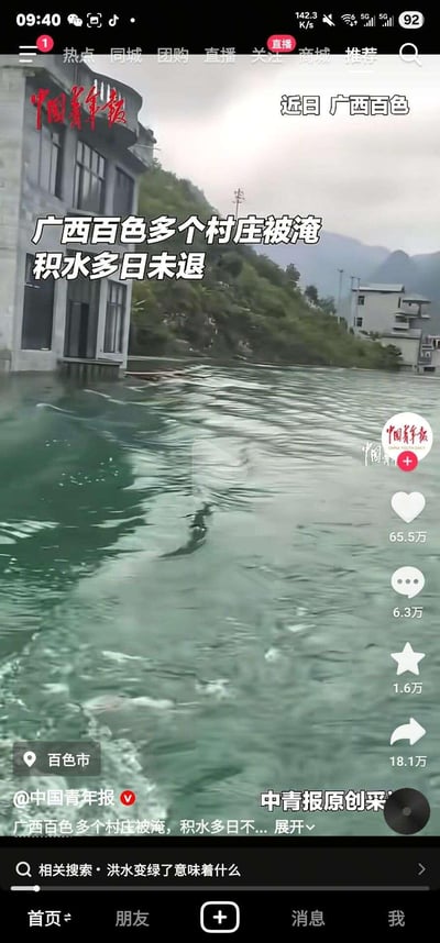 这是中国青年报发布的广西百色洪涝灾害的抖音视频截图，画面中村庄区域被大面积浑浊积水覆盖，水位已淹没至民居低层位置，水面开阔且带有波纹，背景可见山坡与其他被淹房屋，直观呈现出当地多个村庄受灾、积水多日未退的严重洪涝灾情，无谐音梗、双关或暗示类内容。