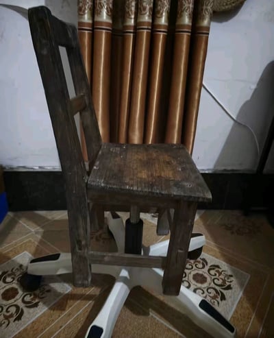 这张图展示了一个风格反差极强的DIY椅子：椅子上部是用了很久、表面磨损掉色的传统实木小板凳，下部却是现代升降转椅的白色带滚轮五星脚底座，两种完全不匹配的部件被组合在一起，充满了违和又接地气的趣味感。背景是带有花纹的棕色窗帘、瓷砖地面，是典型的国内县城或乡村老家的居家场景，契合大学生放假回老家遇到的充满朴实实用主义的生活细节的主题。