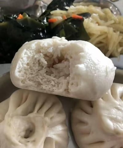 图片呈现了一桌家常餐食，前景是三个白面包子，其中一个被掰开，露出的馅料并非包子常见的肉、菜馅，而是满满白米饭，这个奇特的搭配显得格外突兀。背景里还有两盘配菜，一盘是点缀着橙色胡萝卜丁的深色海带，另一盘是浅黄色的炒土豆丝，整体场景充满生活感，这个米饭馅的包子大概率是制作时的失误，让人觉得既搞笑又无奈。