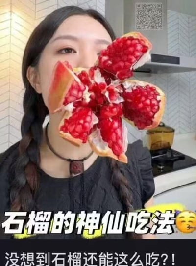 图片中一名女性在厨房环境中，将一个剥开的石榴掰成类似花朵的形状举在脸前，石榴籽鲜红饱满，视觉效果突出。图片底部配有文字“石榴的神仙吃法😋”，旨在展示一种创意的石榴食用或摆盘方式。背景可见带有几何图案的瓷砖墙面及厨房灶台，右上角有一个二维码标识。