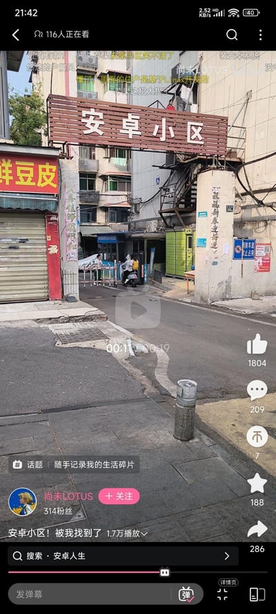 这是B站用户发布的实拍视频截图，画面展示了一个名为“安卓小区”的真实老旧居民小区入口，入口通道旁有“鲜豆皮”店铺，还有骑电动车的居民正常进出。弹幕里的网友玩起了数码圈的关联梗，将小区名字和安卓系统结合，调侃“里面的住户是基于Linux开发的”“苹果这招太狠了”，因为安卓系统本身基于Linux内核开发，用数码知识玩小区名字的梗，十分搞笑。