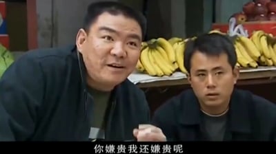 这是电视剧《征服》经典买瓜桥段的截图，画面左侧是卖瓜摊主，身体前倾，面露略带不耐的挑衅神情，右手抬起正在说话，他身旁坐着另一名男子，面露惊讶疑惑的神色，二人后方是摆放着成串香蕉和其他水果的摊位，画面底部配有白色字幕“你嫌贵我还嫌贵呢”，是该名场面中摊主回应刘华强的台词内容。