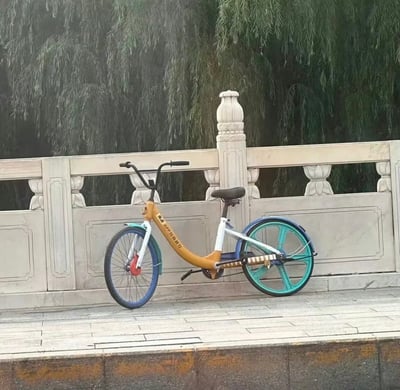 一张户外场景照片，展示了一辆美团共享单车停靠在石栏杆旁。自行车采用黄色车架，搭配蓝色前轮和青绿色后轮，车把和座椅为黑色。背景是茂密的柳树枝叶，石栏杆上有莲花状装饰柱。整体画面呈现出日常城市生活的平静氛围。