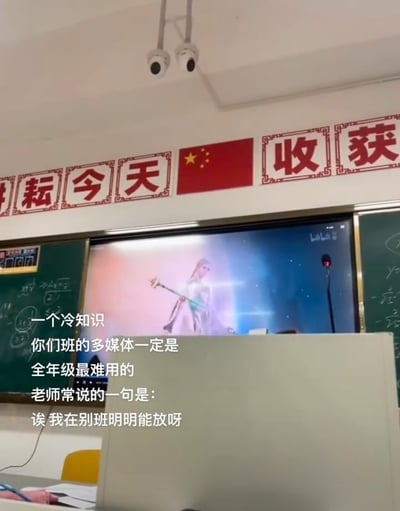 教室场景图，前方投影屏幕显示一位持武器的女性角色，背景墙上悬挂红色标语“耕耘今天”和中国国旗，天花板装有监控。图片叠加文字内容：“一个冷知识 你们班的多媒体一定是全年级最难用的 老师常说的一句是：诶我在别班明明能放呀”，幽默吐槽校园中多媒体设备常见的使用问题，引发学生共鸣。