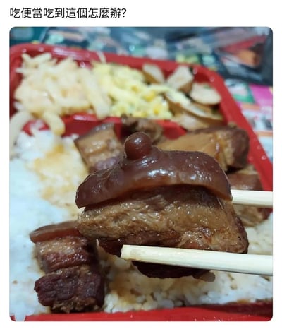 图片展示了一份红色便当盒内的餐食，包含米饭、红烧肉、土豆丝、玉米炒蛋等菜品。画面中心用筷子夹起一块红烧肉，肉的顶部有一个明显的圆形凸起物，形状较为奇特。图片上方配有文字“吃便當吃到這個怎麼辦?”，表达了对便当内出现异常肉块的疑惑。