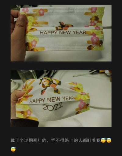 这是一组上下拼接的静态对比图，上图展示了一只手拿着未展开的新年主题口罩，口罩表面印有樱花、日式折扇图案和“HAPPY NEW YEAR”字样；下图是该口罩被展开后的样子，露出了隐藏的“2022”字样。图片下方配有文字“戴了个过期两年的，怪不得路上的人都盯着我😇😇😇”，结合时间来看，发布时应为2024年，这款2022年的新年口罩在当下属于“过时”的主题款式，博主因此遭遇社死，整个场景充满了尴尬又好笑的生活趣味。