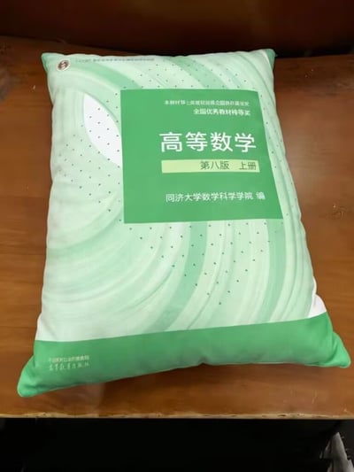 图片中展示了一个印有经典《高等数学（第八版 上册）》教材封面图案的抱枕。抱枕主体为浅绿色，表面印有教材封面的标志性绿色方块，内含白色文字“高等数学 第八版 上册 同济大学数学科学学院 编”及“全国优秀教材特等奖”等字样，背景则是螺旋状的淡绿色波浪纹与点状图案。抱枕被放置在木质桌面上，整体设计将令无数大学生“头疼”的高数教材转化为日常用品，充满校园怀旧感与实用幽默，很可能是针对大学生群体的创意周边产品。