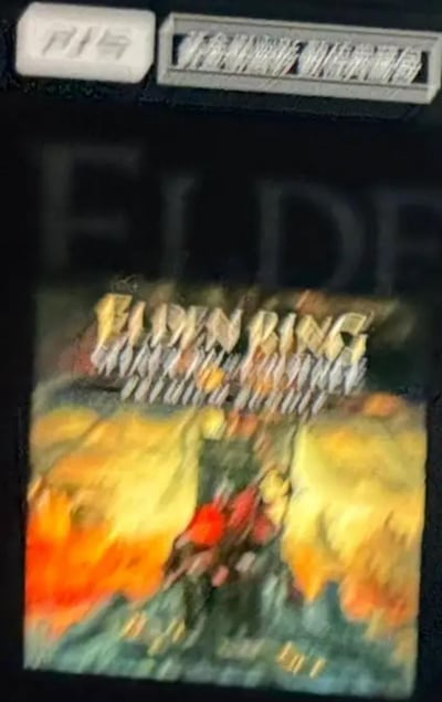 这是一张非常模糊的图片，似乎是艾尔登法环(Elden Ring)游戏封面的照片。图片存在明显的运动模糊，导致细节难以辨认。可以看到标题"ELDEN RING"以金色字体显示在封面上方，下方似乎有游戏场景的艺术画，但因模糊无法看清具体内容。图片顶部还有一些模糊的白色文字或标识，但无法辨认具体内容。