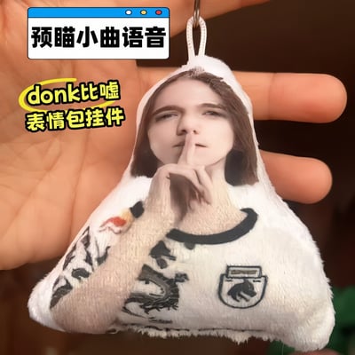 整张图为实拍内容，一只手捏着一个三角造型的毛绒挂件，挂件表面印有CS2职业选手donk做出比嘘噤声动作的经典表情包形象，挂件顶部配有挂绳可用于悬挂。图中添加了两处文字标注，顶部标注该挂件具备预瞄小曲语音功能，左侧标注该商品为donk比嘘表情包周边，是面向电竞爱好者的趣味衍生产品。