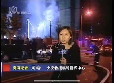这是上海新闻综合频道的见习记者周瑜在火灾救援临时指挥中心进行现场报道的画面。夜晚的现场背景中可见冒烟的建筑、消防车、救援人员及相关车辆，记者手持带有STV标识的话筒，正严肃地进行事件报道，画面下方字幕标注了记者身份与报道地点。画面无谐音梗或双关含义。