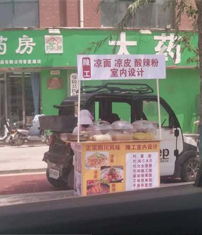 这是一张街头实拍梗图，画面主体是停在绿色招牌药房门前的小型代步车改装的流动小吃摊，摊主戴着粉色遮阳帽在摊位后值守。摊位的宣传内容呈现极强的反差感：既售卖四川风味的凉面、凉皮、酸辣粉等小吃，同时还承接量房、画CAD、做效果图等室内设计相关业务，两种完全不相关的经营内容放在同一个流动摊位上，形成强烈的荒诞笑点。背景可见街边停放的电动车和药房的店面。