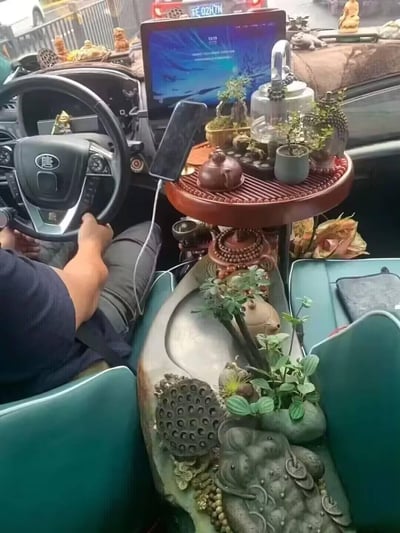 这张照片展示了一辆比亚迪唐汽车的独特内饰：驾驶员正手握方向盘，仪表盘区域放置着一台亮屏笔记本电脑，旁边插着充电的手机。中控台上搭起一个圆形小桌，摆满了茶具（紫砂壶、玻璃茶壶）、多盆迷你绿植和小型雕像，前方挡风玻璃下沿也陈列着一排小摆件。中央扶手区域则是一个大型装饰性茶盘，雕刻有莲花座和金蟾造型，搭配鲜活绿植，俨然将车内打造成了一个移动的中式茶室。整体场景充满生活气息与个性创意，展现了车主对茶文化的喜爱和独特的车内空间利用方式。