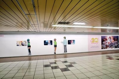 这是一张地铁通道内的照片，墙面展示着《偶像大师》（THE IDOLM@STER）20周年纪念的系列动漫海报，右侧海报明确标注“20周年 THE IDOLM@STER”及日文“アイドルマスター 20周年”字样。墙面上立有三个等身大的角色立牌：左侧为绿衣短发女性角色站立注视海报，中间为绿衣女性角色自然站立，右侧为浅蓝色上衣男性角色伸手指向海报。地面是黑白格子地砖，天花板为木质条状结构并装有长条形灯管，整体环境明亮整洁，营造出浓厚的动漫周年纪念氛围。