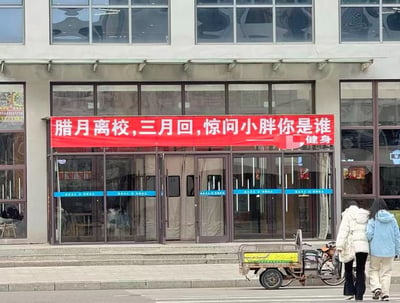 这是一张实拍校园周边健身馆门口的照片，建筑入口处悬挂着一条醒目的红色横幅，上面写着「腊月离校，三月回，惊问小胖你是谁」，用贴近学生寒假离校、春节后返校体重增长的生活场景玩梗，以此吸引有减肥需求的学生顾客，横幅右下角标注「健身」点明店铺业务。画面下方有两位穿着冬装的女生正从门前走过，路边还停着一辆三轮车，整体场景真实接地气，容易引发学生群体的共鸣。