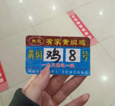 这是一张线下餐饮门店取餐号牌的实拍图，一只带有美甲的手拿着该号牌。号牌顶部为红底黄字，标注“朱記 有家黄焖鸡”；中间蓝底区域，黄字“黄焖”与“号”之间，用白色方块分别手写“鸡”和“8”，组成“黄焖鸡8号”，但“鸡”与“8”的组合形成了不雅谐音梗；下方配有红底白字标语“一生只做这一锅”，最底部白字写着服务标语“味道好请告诉别人 不好请告诉我们”。号牌表面有明显的使用褶皱痕迹，背景是浅色瓷砖地面，能看到部分红色标识。