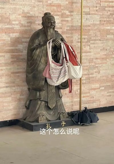 图片中是一尊孔子的雕像，被当作包架使用，雕像手臂上挂满了几个手提包，脚下还有雨伞放置，画面明显带有日常生活与尊重历史人物形象的反差。图片配有"这个怎么说呢"的幽默文字，传递出无奈又好笑的气氛。文字与孔子的形象产生了文化意义上的反差和调侃，暗含双关和吐槽现实的不合适。