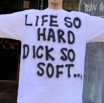 一张展示白色T恤背面的照片，T恤上用黑色手写体写着英文短语：'LIFE SO HARD DICK SO SOFT...'。图中人物双臂张开，背景是带有涂鸦的城市街道墙面。通过生活艰难与身体状况的对比制造黑色幽默效果。