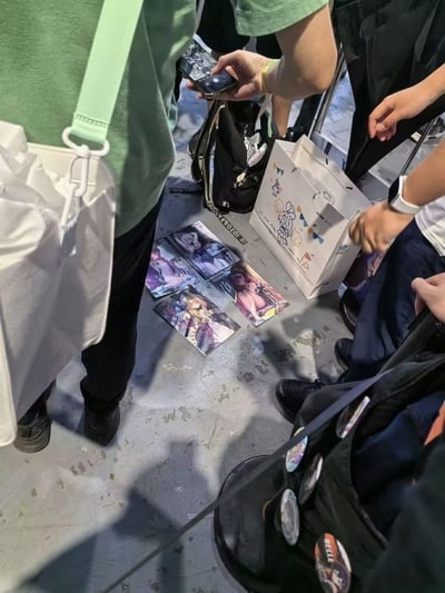 这是一张在ACG漫展现场拍摄的照片，地面上摆放着四张风格各异的二次元美少女收藏卡片，周围聚集了几位ACG爱好者，有人手持手机，有人携带周边背包，看起来正在围观或进行卡片的交流、交易，是线下ACG爱好者社群互动的典型场景。