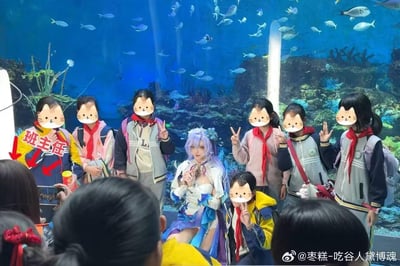 这是一张在水族馆内拍摄的现场照片，画面中央是一位cosplay《原神》神里绫华的女性，她被一群系着红领巾的小学生围绕着，其中一位被红色箭头和文字标注为“班主任”的老师也在队伍中。孩子们对着镜头比出剪刀手，显得十分兴奋，coser则端坐做出温婉的手部动作。背景是大型水族箱，里面有游动的鱼类和珊瑚造景，前景还有其他围观的人群，整体氛围轻松愉快，充满了次元文化与日常校园场景碰撞的趣味。