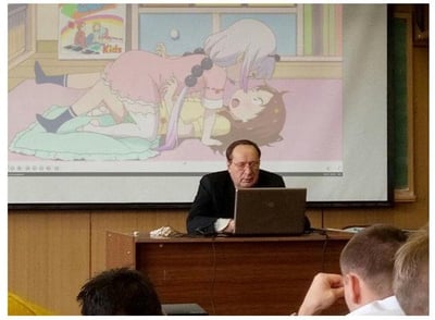 这是一张在课堂/讲座现场的拍摄图，一位年长男性讲师坐在讲台后操作笔记本电脑，身后的投影幕布却播放着日本动画《小林家的龙女仆》中的经典场景：康娜卡姆依压在才川莉子身上，严肃的线下教学场景与搞笑的二次元动画内容形成强烈反差，制造出滑稽的喜剧效果，图中没有双关、谐音梗或暗示内容。