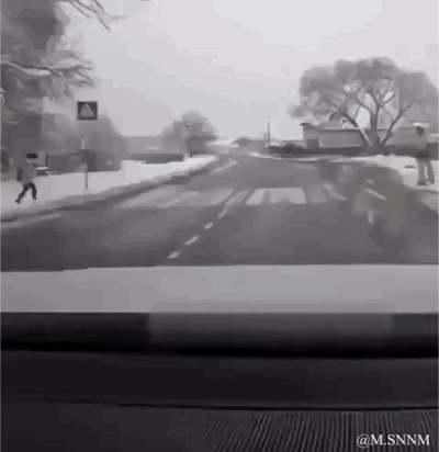 这是一段从车内视角拍摄的动态GIF，场景是积雪覆盖的冬季乡村道路，道路两侧有行人。当车辆向前行驶时，右侧路边的行人与路边的标牌恰好形成视觉错位，看起来像是行人被车辆撞到，实际是视觉错觉，制造出戏剧性的搞笑效果，让人看完后会心一笑。