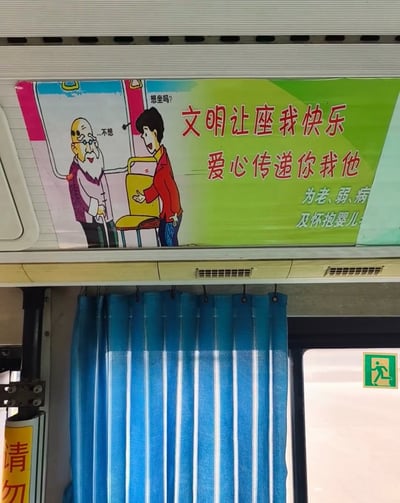 这是一张拍摄于公交车内部的实拍照片，车厢顶部张贴着文明让座主题的宣传海报：海报右侧是红色宣传语「文明让座我快乐 爱心传递你我他」，下方小字标注让座服务对象为老、弱、病及怀抱婴儿的群体；海报左侧是卡通插画，穿红衣的年轻乘客笑着向拄拐杖的白发老人邀座，旁配文字「想坐吗？」，但老人的心理活动配文为「...不想」，和常规公益宣传的正向逻辑形成反差，产生搞笑效果。海报下方可见车厢内的蓝白竖条纹窗帘、车窗旁的绿色安全出口标识，左侧还露出了黄色的「请勿」开头的提示标识边缘。