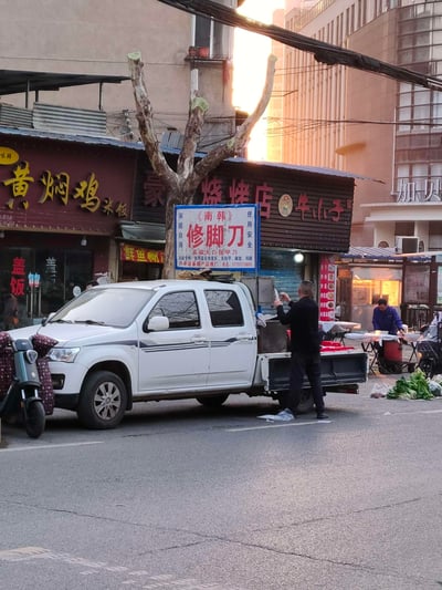 城市街道旁的日常景象，有多家小吃店包括黄焖鸡米饭和烧烤店。一辆白色皮卡车停在路边，一名男子正在车旁准备食物。最引人注目的是一个写着《南韩》修脚刀的招牌，与周围的餐饮店铺形成有趣对比。背景中还有其他摊贩和行人，展现了典型的中国城市街头生活。