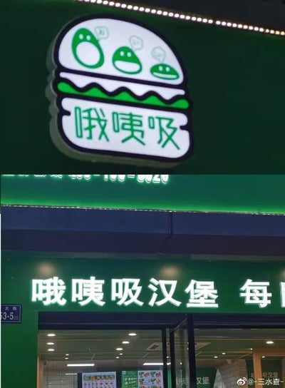 这是一张线下汉堡店的实拍竖图，展示了店铺的招牌与门脸区域。上方的招牌为创意汉堡造型，内部包含三个圆润的绿色卡通形象，下方配有店铺名称“哦咦吸”；下方门脸处的发光白色字体写着“哦咦吸汉堡 每”，后续文字被画面截断，左下角可见门牌号“3-5”。店名“哦咦吸”是谐音拟声梗，模仿人们享用汉堡时发出的进食拟声词，生动有趣，能快速让顾客联想到吃汉堡的满足感，增强品牌记忆点。