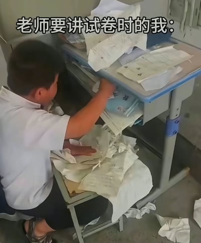 图片中一名穿着白色校服的小男孩正跪坐在教室的课桌旁，焦急地翻找着抽屉和散落在地上、桌面上的试卷。课桌内外堆满了各种纸张，显得十分凌乱。图片上方配有文字“老师要讲试卷时的我：”，幽默地描绘了学生在老师即将讲解试卷时，因找不到自己的卷子而手忙脚乱的场景，生动还原了校园生活中常见的慌张时刻。