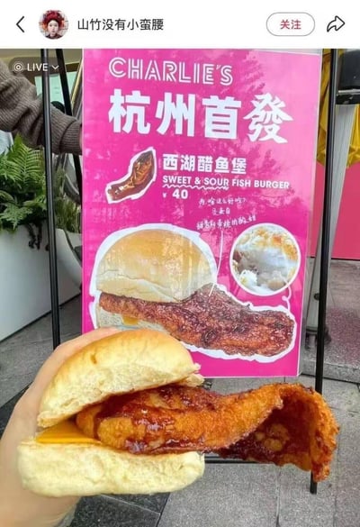 社交媒体用户拍摄的杭州某餐厅新品宣传现场，画面中一只手正拿着刚购买的西湖醋鱼堡，前方立着粉色的CHARLIE'S餐厅广告牌，展示着同款汉堡的宣传图。广告牌上写着"杭州首發 西湖醋鱼堡"，售价40元，并配有产品特写图片和"为啥这么好吃？是来自甜密和带挑的酸文相"的宣传语。实物汉堡中的鱼排看起来比宣传图更大，形状更不规则。