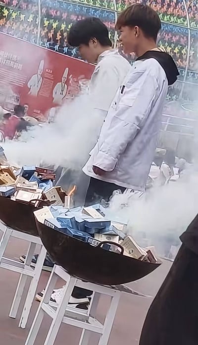这是一张实拍照片，画面里两口大铁锅被架在白色高脚凳上，锅内堆满了各式各样的香烟盒，正在焚烧产生大量白色烟雾，旁边站着数名穿着浅色外套的年轻男性，疑似在校学生，正在观看香烟焚烧的过程。该场景被网友类比为历史事件虎门销烟，搭配的文案玩了双关梗，将香烟品牌“利群”谐音为“利于群体”，调侃“一个人抽是利己 一群人享受才是利群啊”。