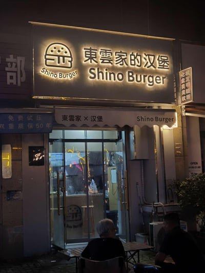 这是一张夜晚拍摄的街边汉堡店实拍图，主角是名为“東雲家的汉堡（Shino Burger）”的独立小店，门店招牌为发光设计，搭配汉堡图标与中日英结合的店名，风格简约温馨。店门口摆放着小桌椅，有两位顾客坐在桌旁，周围还能看到其他店铺的招牌，整体呈现出日常的夜间街边美食小店氛围，没有双关、谐音梗等特殊内容。