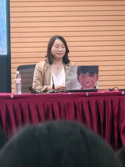 一位女性演讲者坐在铺着红色桌布的会议桌后，面前放着一台笔记本电脑。她穿着米色西装外套和白色内搭，面带微笑，面前有麦克风。笔记本屏幕上显示着一张表情夸张的魔性人脸图片，形成了正式场合与搞笑内容的有趣反差。桌子上还放着一瓶矿泉水和一副眼镜。背景是木质墙面，前景能看到部分观众的头顶。