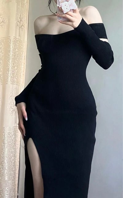 一位女性在室内环境中自拍，她穿着一件黑色露肩长袖连衣裙，裙子采用修身针织材质，侧面有高开叉设计。她左手拿着手机自拍，右手自然下垂放在大腿旁，背景中可以看到带有花纹的浅色窗帘。整体穿搭展现出优雅性感的风格，适合日常穿着或特殊场合。