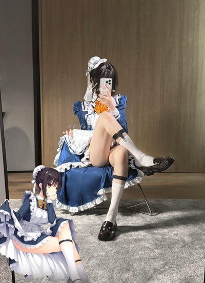 图中呈现了三次元与二次元同框的趣味场景：一位穿着蓝白女仆装的真人coser坐在椅子上，举着手机对镜自拍，她的女仆装造型（包括蓝白裙装、白色头饰、袜圈、制服鞋等细节）高度还原了左下角的二次元女仆角色，该二次元角色正托腮坐在地面，神态慵懒。两者搭配形成了软萌可爱的视觉效果。
