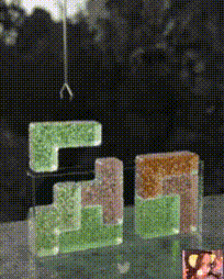 这是一则3D渲染的动态GIF图，画面中由绿色、棕色的像素风格3D方块组合成近似谷歌（Google）品牌标识的造型，画面上方设有一个金属吊钩，吊钩会吊起其中一块方块并移动，实现方块的重组效果，背景为模糊的深色场景，右下角附带一个小尺寸的缩略图，整体通过动态的吊装动作，将静态的品牌标识转化为具有互动感的视觉内容，充满创意趣味。