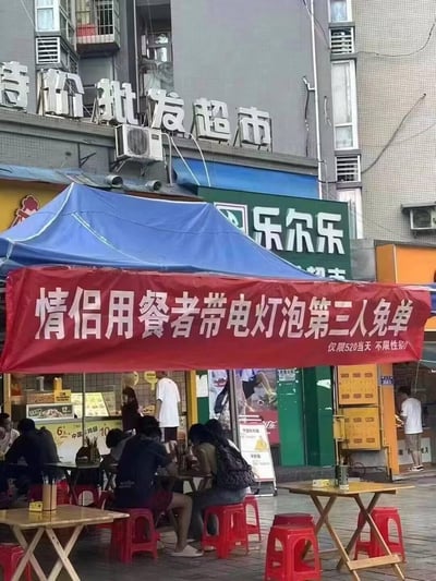 街头一家名为'乐尔乐'的店铺外，搭着蓝色帐篷，帐篷下挂着红色促销横幅，上面写着'情侣用餐者带灯泡第三人免单 仅限520当天 不限性别'。帐篷下摆放着几张桌子和红色塑料凳子，有几桌人正在用餐。背景中可以看到'特价批发超市'的招牌和居民楼。

这里的'灯泡'是网络流行语，指的是在情侣约会时陪伴在旁的单身人士，即'电灯泡'，店家巧妙运用这个梗推出促销活动，吸引顾客。