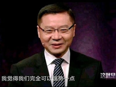 图片中是一位戴着眼镜、穿着黑色西装和条纹领带的中年男性，他面带微笑，正在进行发言。背景为深色带有紫色调的模糊布景，人物胸前佩戴有麦克风。图片底部有白色文字：“我觉得我们完全可以强势一点”，右下角有部分文字“这就是”。