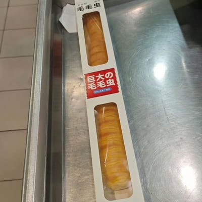 图片是一根长条面包，包装上的开窗部分展示出面包本身，由于造型和表面纹理，很像一条毛毛虫。包装纸上的红色日文标签写着“巨大的毛毛虫”，让商品在人们认知中形成食品与生物的双关趣味。这种梗图常用于吐槽食品造型或日式脑洞包装。

双关点：文字与形象形成了“真买了毛毛虫”的视觉和语义幽默。