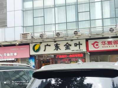 这是一张街头实拍的日常城市街景照片，前景是两辆汽车的车顶，背景是一栋带有玻璃幕墙的多层建筑底层商铺区域。从左至右可见三家店铺：粉色招牌的克丽缇娜美容店、白底黑字带品牌标识的广东金叶（赤岗专营店）、红白色的华夏银行网点，建筑外墙上安装有多台空调外机，整体场景平实普通，记录了城市里常见的商铺街面样貌，无谐音梗或双关含义。