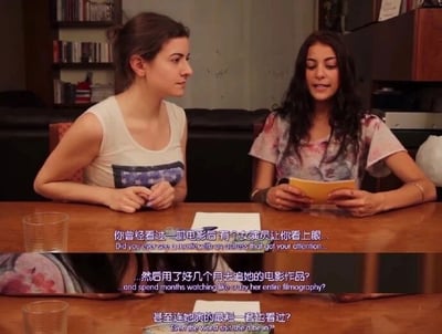 这是一张影视截图，画面呈现了室内场景中两位年轻女性坐在木质餐桌前的互动画面：左侧女生穿着带有美国国旗图案的白色无袖上衣，侧头专注地看向右侧女生，双手交握放在桌上；右侧女生穿着晕染风格的短袖上衣，手里拿着一个黄色本子，正开口说话。画面下方配有中英双语字幕，中文内容为“你曾经看过一部电影后有个女演员让你看上眼...然后用了好几个月去追她的电影作品？甚至连她演的最烂一套也看过？”，这段对话围绕影迷追星补片的经历展开，很容易引发影迷群体的共鸣。对于图中的问题，很多影迷都有过类似经历：当被某一位演员的演技或者个人魅力吸引后，会花费大量时间去观看该演员的所有影视作品，哪怕是评价不佳的作品，只为了更完整地感受该演员的成长和不同的表演风格。