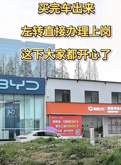 整张图为实拍场景，左侧是比亚迪品牌的线下汽车销售门店，右侧与其紧邻的是滴滴出行的授权司机招募门店，招募门店外的宣传牌标注有跑滴滴的相关招聘福利内容。图片上方配有黄底黑边的醒目文字，以幽默调侃的方式讽刺了当下消费者购车后可以直接入职成为网约车司机的现象，暗指汽车销售和网约车行业形成了看似双赢的有趣商业闭环。