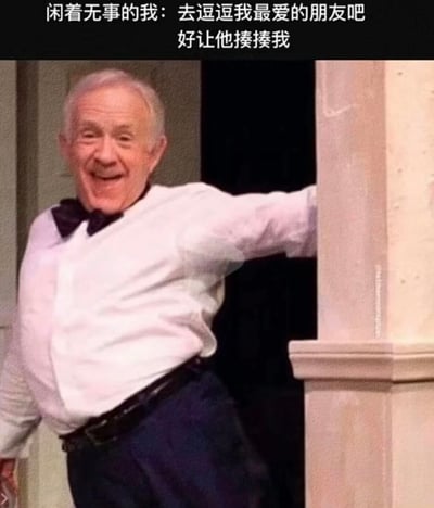 图片中是一位白发老人，穿着白色衬衫和黑色领结，侧身倚在石柱上，脸上带着开心的笑容，身体微微扭曲，姿态显得有些滑稽。图片上方配有文字“闲着无事的我：去逗逗我最爱的朋友吧 好让他揍揍我”，通过老人搞怪的表情和动作，配合文字内容，形成一种幽默效果，展现了朋友之间互相调侃、打打闹闹的亲密关系。