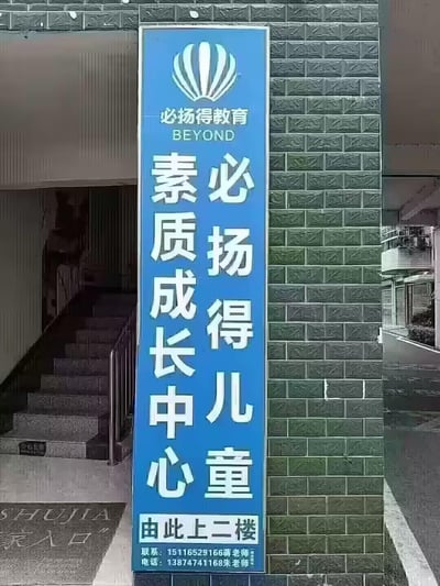 这是一张竖版的蓝色教育机构指引招牌，安装在贴有深绿色纹理瓷砖的建筑墙面上，招牌旁是通往二楼的室内楼梯。招牌顶部配有贝壳状品牌标识，标注“必扬得教育 BEYOND”，中间以竖排双列的形式呈现核心名称“必扬得儿童 素质成长中心”，底部明确指引“由此上二楼”，并附上了蒋老师和朱老师的联系电话，用于引导家长和儿童找到位于二楼的线下素质成长教育机构，整体风格正式清晰，无谐音梗或双关内容。
